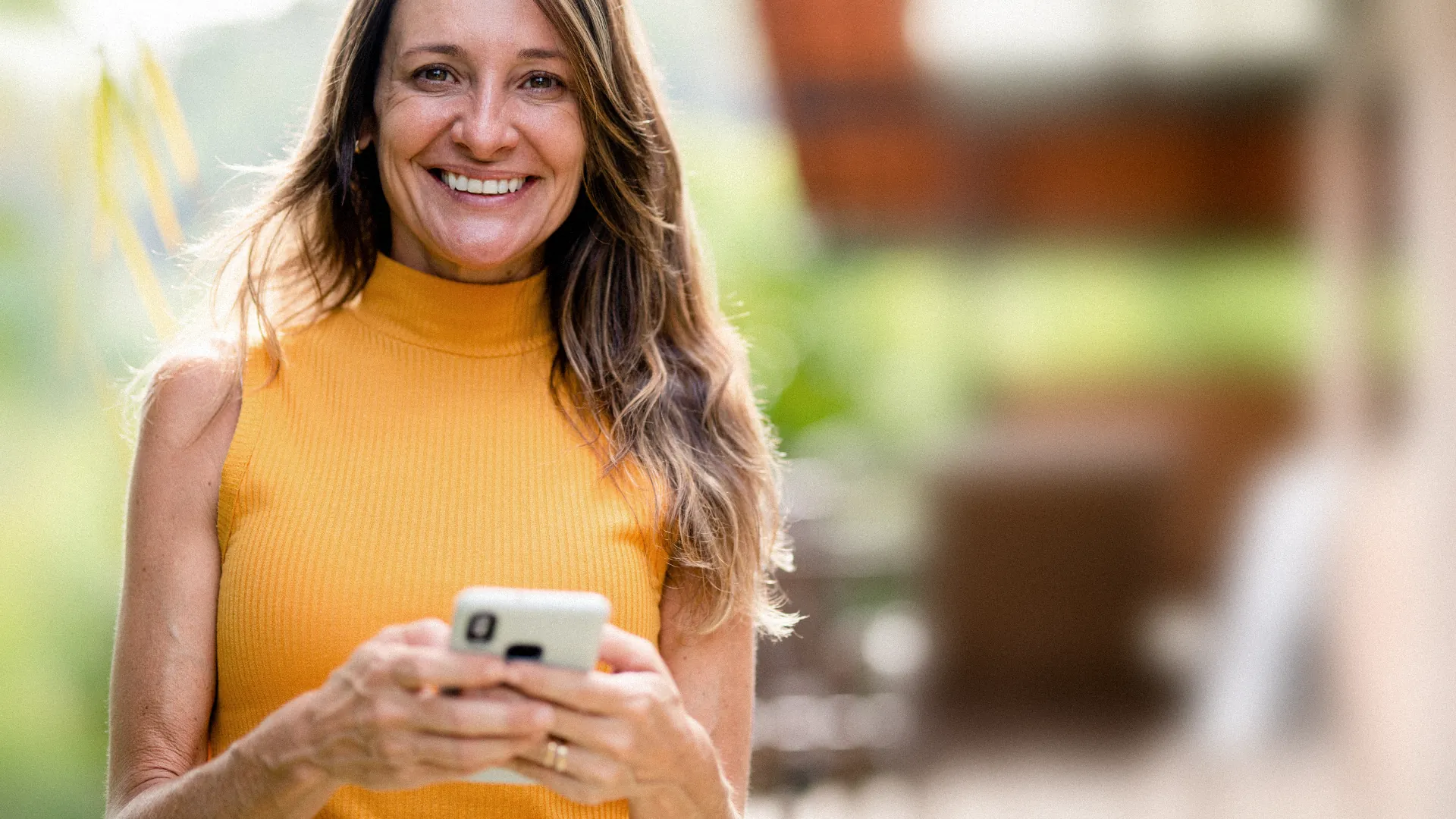 Smiling woman in an orange top holding her phone outdoors, looking straight at the camera