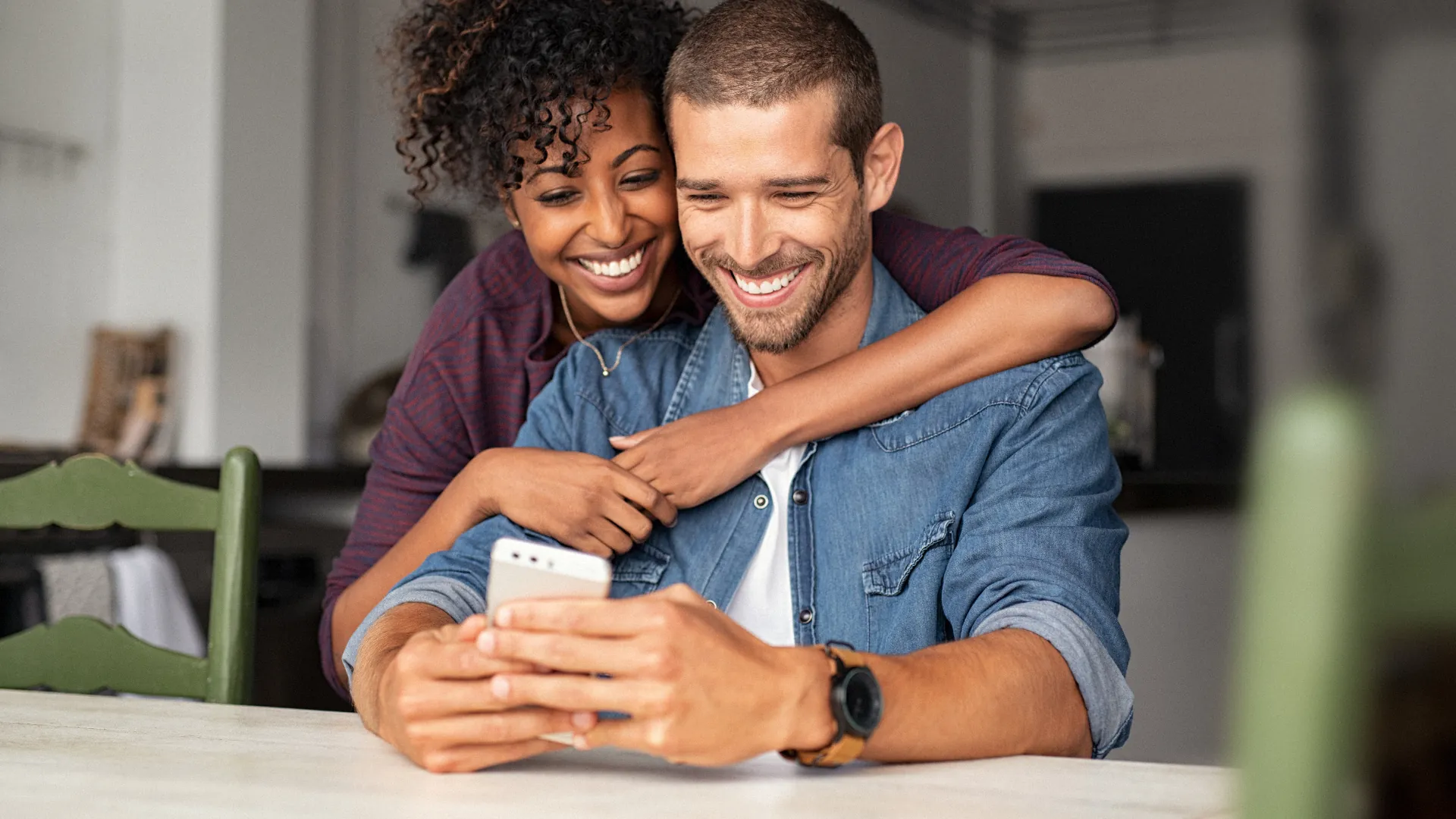 Laughing couple sharing a phone at a kitchen table, her arms wrapped around his shoulders