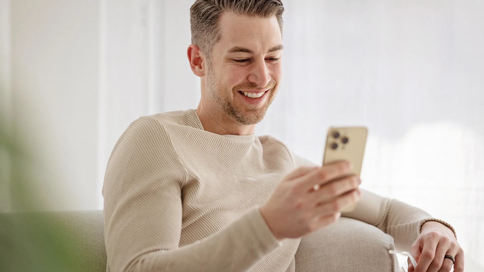 Relaxed man in a cream sweater smiling at his phone on a sofa in a bright room